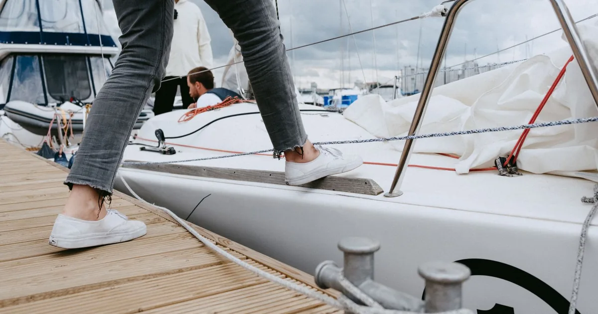 A person steps from the wooden pontoon onto the deck of a sailing yacht, the crew waiting on board