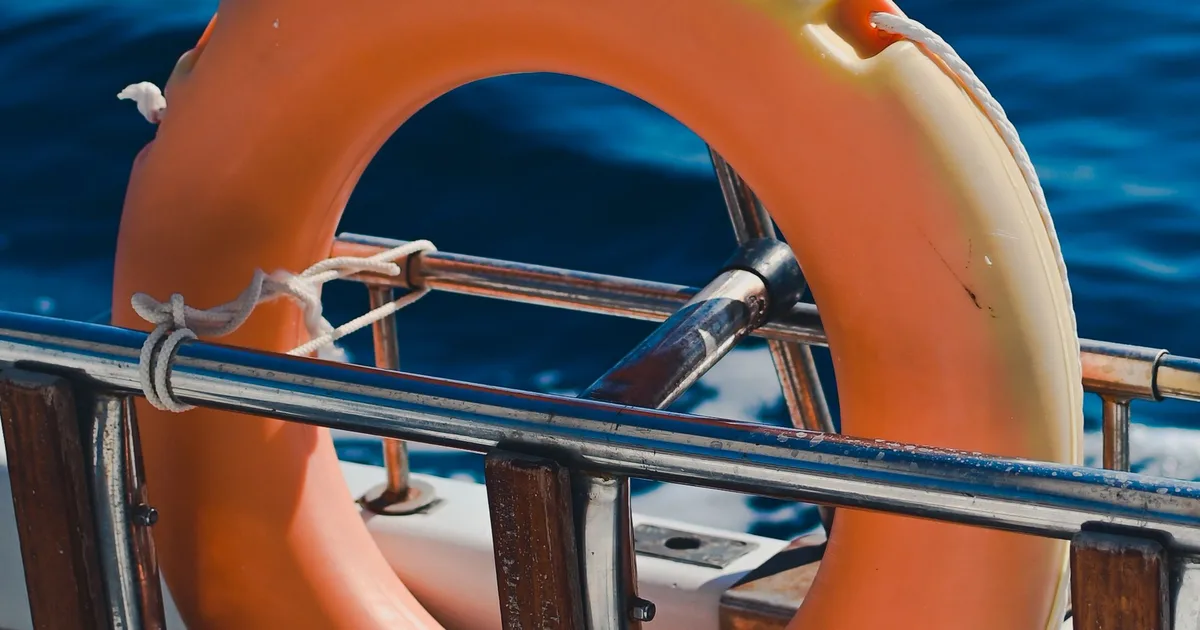 An orange lifebuoy hangs on the guardrail of a sailing yacht, the open sea behind it
