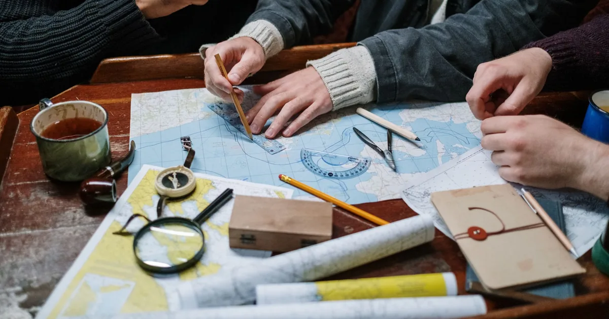 Several hands plotting a route on a nautical chart with a plotter, dividers and pencil, a compass and a mug of tea beside them